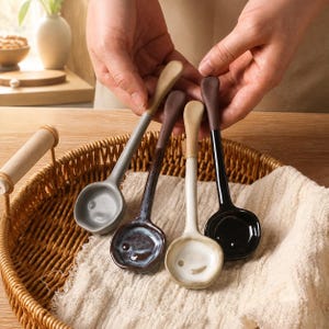 May include: Four ceramic spoons with long handles and rounded bowls. The spoons feature distinct finishes: a matte grey, a dark brown with a mottled texture, a cream-colored bowl with a light brown handle, and a glossy black with a brown handle.