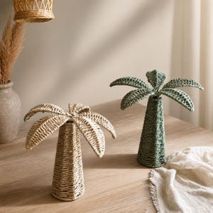 Op de afbeelding: Twee decoratieve palmboomsculpturen gemaakt van gevlochten natuurlijke vezels. De ene sculptuur is lichtbeige van kleur, terwijl de andere een gedempte saliegroene tint heeft. Beide hebben een conische stam en uitwaaierende palmbladeren.