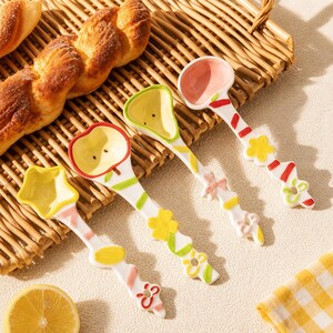 May include: A set of four ceramic spoons with white handles decorated with colorful floral and geometric patterns. The spoon bowls are shaped like a star, an apple, a pear, and a round scoop, featuring pastel pink, yellow, and green interior colors.