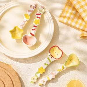 May include: Four ceramic spoons with decorative handles featuring painted bows, flowers, and stripes. Two spoons have star and round bowls, while the other two feature apple and pear-shaped bowls with small dot details in yellow and green.