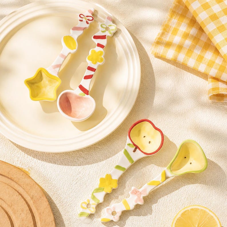 May include: Four ceramic spoons with decorative handles featuring painted bows, flowers, and stripes. Two spoons have star and round bowls, while the other two feature apple and pear-shaped bowls with small dot details in yellow and green.