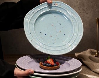 Könnte beinhalten: Ein Stapel runder Keramikteller mit gesprenkelter Oberfläche und konzentrisch gerillten Rändern. Der oberste Teller ist hellblau mit bunten Sprenkeln, der darunter liegende Teller ist lavendelfarben und mit einer Beeren-Tarte belegt.