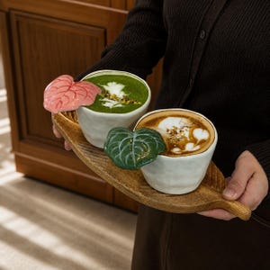 Peut inclure: Deux tasses en céramique blanche aux surfaces irrégulières et texturées, chacune dotée d'une anse en forme de feuille. Une tasse a une anse feuille rose et contient du matcha vert, l'autre a une anse feuille verte et contient du café.
