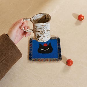 Puede incluir: Un posavasos cuadrado de tejido azul con un gráfico de gato negro con una cereza roja en la cabeza y bordes de flecos multicolores, acompañado de una taza de cerámica veteada en blanco y marrón y dos tomates cherry rojos.