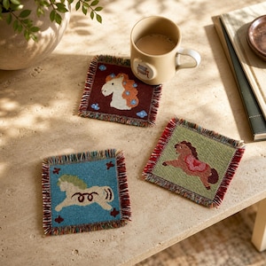May include: Three square woven coasters with fringed edges, each featuring a different horse illustration. One is maroon with a white horse, one is blue with a cream horse, and one is sage green with a brown horse, all with colourful woven details.
