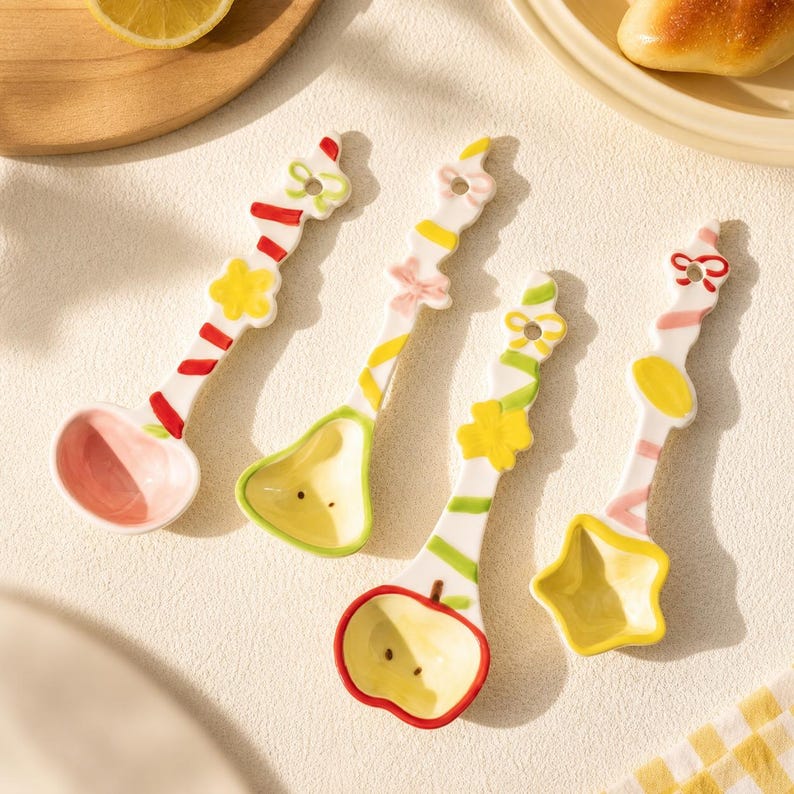 May include: Four ceramic spoons with white handles featuring colorful painted patterns of bows, flowers, and stripes. The spoon bowls are shaped like a circle, a pear, an apple, and a star, each finished in pastel pink, yellow, or cream tones.