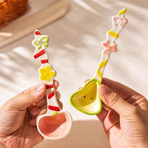 May include: Two ceramic spoons with decorative handles. The left spoon has a pink bowl, a white handle with red stripes, and a yellow flower accent. The right spoon has a yellow bowl, a white handle with yellow stripes, and a pink bow accent.