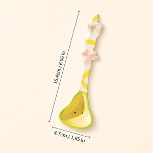 May include: A ceramic spoon featuring a yellow and white striped handle with a pink bow accent and a yellow bowl. The spoon measures 6.06 inches in length and 1.85 inches in width as indicated by the visible measurement lines and text.