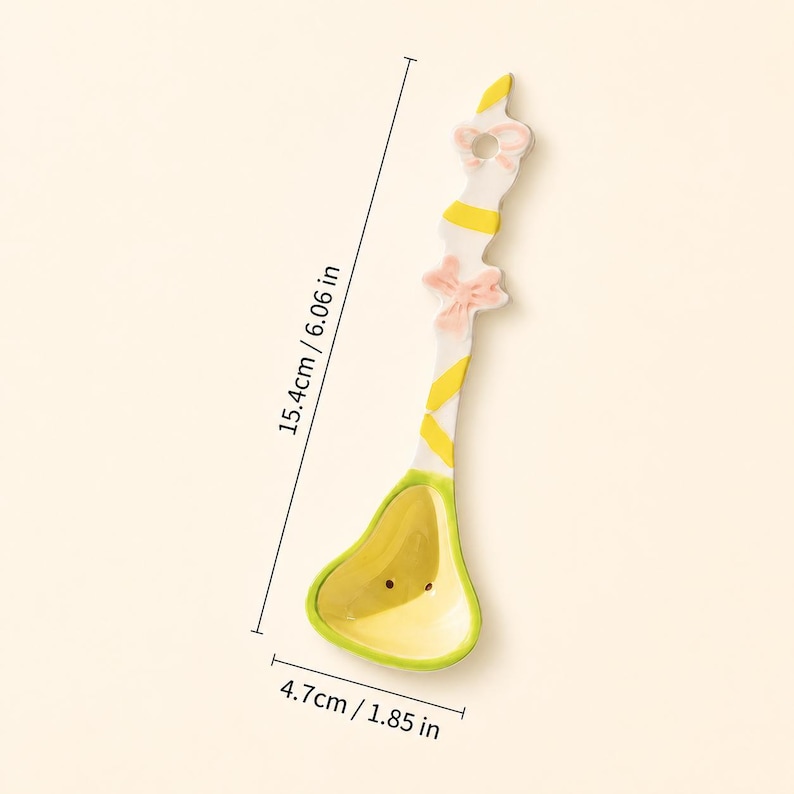 May include: A ceramic spoon featuring a yellow and white striped handle with a pink bow accent and a yellow bowl. The spoon measures 6.06 inches in length and 1.85 inches in width as indicated by the visible measurement lines and text.