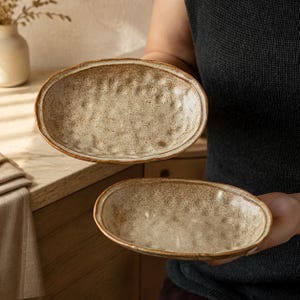 May include: Two oval-shaped ceramic dishes with a speckled, off-white glaze and a textured, dimpled surface. The edges of the dishes feature a darker brown, earthy rim, highlighting the organic, hand-formed shape of the stoneware pottery pieces.
