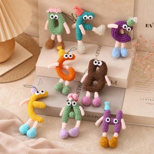Può includere: Una collezione di otto ornamenti a forma di lettere all'uncinetto, ognuno dotato di due grandi occhi bianchi con pupille nere, braccia sottili e gambe penzolanti con piedi rotondi. Le lettere sono realizzate in vari colori di filato, tra cui verde, verde acqua, viola, arancione, marrone, giallo e lavanda, con piccoli cappellini abbinati su alcune.