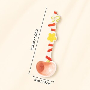 May include: A ceramic spoon with a pink bowl and a white handle featuring red diagonal stripes, a yellow flower, and a light green bow. The spoon measures 6.02 inches in length and 1.97 inches in width, as indicated by the visible measurement lines.