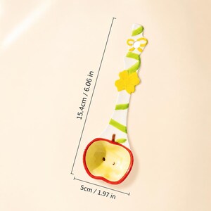 May include: A white ceramic spoon with a red and yellow apple-shaped bowl, featuring a green vine pattern, a yellow flower, and a bow on the handle. The spoon measures 6.06 inches in length and 1.97 inches in width, as indicated by the text.