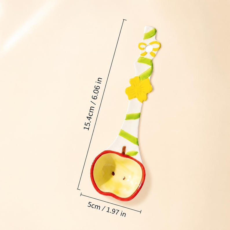 May include: A white ceramic spoon with a red and yellow apple-shaped bowl, featuring a green vine pattern, a yellow flower, and a bow on the handle. The spoon measures 6.06 inches in length and 1.97 inches in width, as indicated by the text.