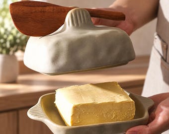 Könnte beinhalten: Eine Keramik-Butterdose mit strukturierter, hellgrauer Oberfläche und passendem Deckel mit Schlitz für ein hölzernes Buttermesser. Der Boden enthält ein rechteckiges Stück gelbe Butter, der Deckel ist angehoben, um den Inhalt zu zeigen.
