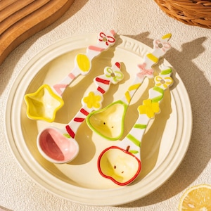 May include: Three ceramic spoons with decorative handles featuring colorful painted patterns of bows, stars, and stripes. The spoon bowls are shaped like a star, a pear, and an apple, with pastel pink, yellow, and red accents on a white base.