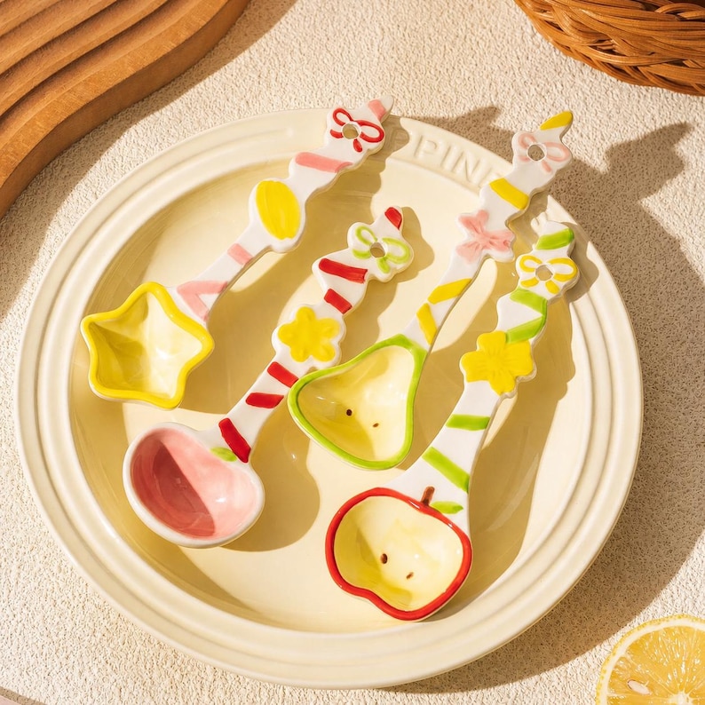 May include: Three ceramic spoons with decorative handles featuring colorful painted patterns of bows, stars, and stripes. The spoon bowls are shaped like a star, a pear, and an apple, with pastel pink, yellow, and red accents on a white base.