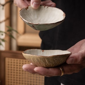 May include: Two small, shallow ceramic bowls with an irregular, organic shape. The interior of each bowl is a smooth, off-white glaze, while the exterior features a textured, earthy brown finish. The rims are uneven with a rustic, metallic-toned edge.