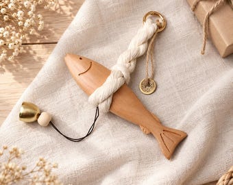 Könnte beinhalten: Ein hölzernes Fischornament mit glatter, hellbrauner Oberfläche und eingekerbtem Lächeln. Es ist an einer dicken, cremeweißen Kordel befestigt, die einen goldfarbenen Metallring, eine kleine Holzperle, eine Messingglocke und einen runden Anhänger trägt.