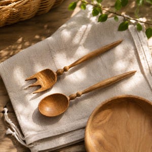 Könnte beinhalten: Eine Holzgabel und ein Holzlöffel mit langen, schlanken Griffen, die ein kleines kugelförmiges Detail am Hals aufweisen. Die Utensilien sind aus hellbraunem Holz mit sichtbarer Maserung gefertigt und liegen diagonal auf einem Stofftuch.