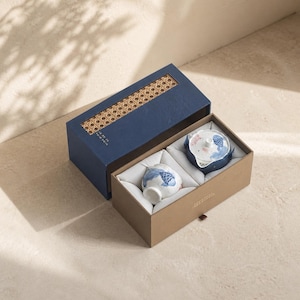 May include: A tea set in a two-part box with a blue lid featuring a woven cane panel and a tan base. Inside, a white ceramic teapot and a matching cup are nestled in white fabric, both decorated with blue and pink koi fish illustrations.