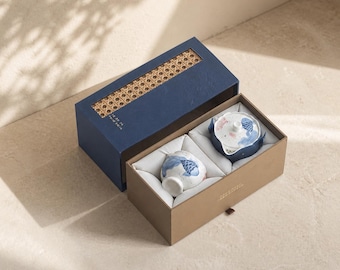 Könnte beinhalten: Ein Teeservice in einer zweiteiligen Box mit blauem Deckel, der ein geflochtenes Rattan-Element aufweist, und einem beigen Unterteil. Darin befinden sich eine weiße Keramik-Teekanne und eine Tasse, eingebettet in weißen Stoff, mit blau-rosa Koi-Motiven.