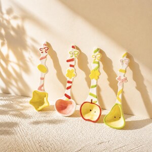 May include: Four ceramic spoons with white handles featuring colorful painted stripes and small decorative shapes at the top. The spoon bowls are shaped like a yellow star, a pink circle, a red-rimmed apple, and a light green pear-like form.
