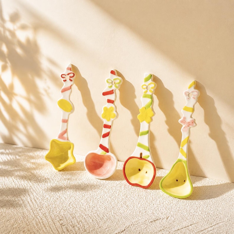 May include: Four ceramic spoons with white handles featuring colorful painted stripes and small decorative shapes at the top. The spoon bowls are shaped like a yellow star, a pink circle, a red-rimmed apple, and a light green pear-like form.
