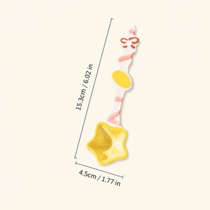 May include: A ceramic spoon with a star-shaped bowl in yellow and a white handle featuring pink stripes, a yellow oval accent, and a red bow detail at the top. The spoon measures 6.02 inches in length and 1.77 inches in width at the star bowl.