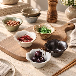 Könnte beinhalten: Eine Sammlung kleiner, blumenförmiger Keramikschalen in verschiedenen Farben wie Salbeigrün, Creme, gesprenkeltem Beige und Dunkelbraun. Die Schalen sind auf einem hölzernen Servierbrett angeordnet und enthalten teilweise Lebensmittel wie Nüsse.