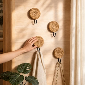 Puede incluir: Cuatro ganchos de pared circulares con una base de ratán tejido natural y un gancho de metal negro. Los ganchos están montados sobre una superficie de madera clara, uno sostiene un paño beige y otro una correa de cadena dorada.