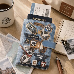 May include: A blue denim passport holder featuring various cat-themed stickers, including illustrations of Siamese cats, a blue crayon, and text reading "Rich," "Miaomiao," "Perfect Day," and "Cat." The holder has a blue snap closure and contains a passport.
