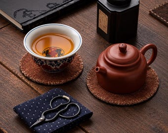 Könnte beinhalten: Eine Keramik-Teetasse mit blau-rotem Blumenmuster und bernsteinfarbener Flüssigkeit, eine Terrakotta-Teekanne, zwei runde, braune, strukturierte Untersetzer, eine schwarze Teedose und eine kleine schwarze Metallschere auf einem dunkelblauen Stoff mit Punkten.