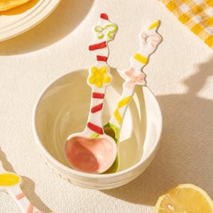 May include: Two ceramic spoons with white handles featuring colorful painted patterns of red stripes, yellow flowers, and green bows, resting inside a plain white ceramic bowl. The spoon bowls are painted in a soft pastel pink and light green.
