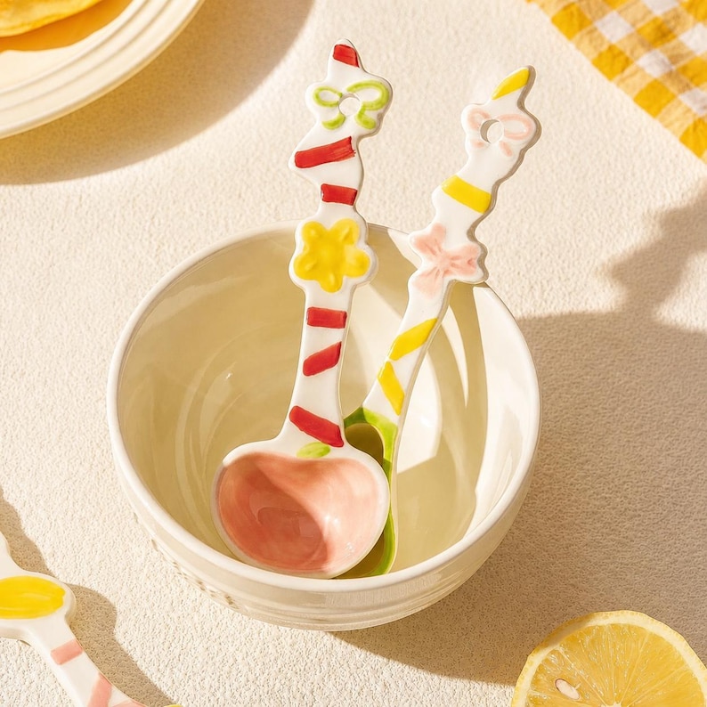May include: Two ceramic spoons with white handles featuring colorful painted patterns of red stripes, yellow flowers, and green bows, resting inside a plain white ceramic bowl. The spoon bowls are painted in a soft pastel pink and light green.