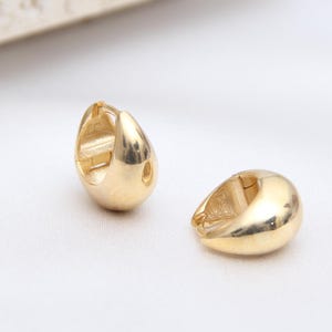 May include: A pair of small, polished gold-tone hoop earrings with a thick, rounded, teardrop-like shape. The earrings feature a smooth, reflective metallic surface and a hinged closure mechanism, presented as two individual pieces on a white surface.