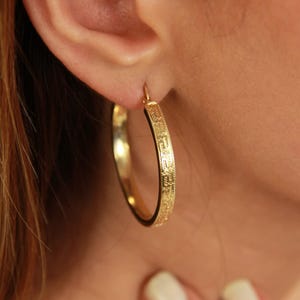 May include: A gold hoop earring featuring a textured Greek key pattern along the outer edge. The circular earring is polished with a reflective finish and is secured through the earlobe with a thin metal wire closure mechanism.