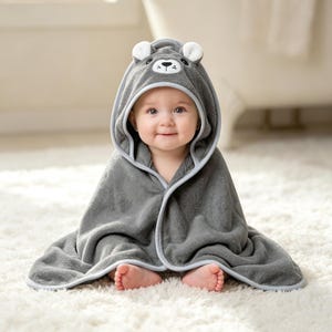 May include: A soft, dark gray hooded towel featuring a bear face design on the hood with white-tipped ears, black embroidered eyes, and a nose. The towel is made of plush fabric with light gray trim along the edges, providing full coverage.