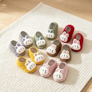以下が含まれることがあります： 赤、黄、緑、グレー、茶、ピンクなど、様々な色のふわふわしたローカットスリッパ10足セット。各スリッパの前面には、ピンクの耳と刺繍のディテールが施された、白いウサギの顔のぬいぐるみアップリケが付いています。