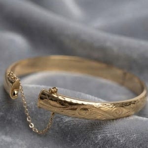 Pode incluir: Uma pulseira rígida articulada de tom dourado com um intrincado padrão de volutas gravado na sua superfície. A pulseira inclui um mecanismo de fecho seguro e uma delicada corrente de segurança fina que liga ambas as extremidades do aro.