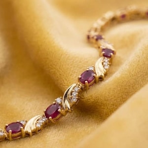 May include: A gold link bracelet featuring a repeating pattern of oval-cut deep red rubies and small clear round-cut diamonds. The metal links are curved and polished, connecting each gemstone setting in a continuous, flexible chain design.