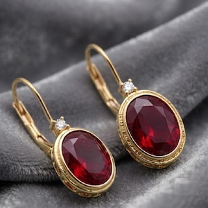May include: A pair of gold leverback earrings featuring oval-cut deep red gemstones. Each earring is accented with a small, clear round-cut stone at the top of the setting, with intricate metal detailing around the oval gemstone bezel.