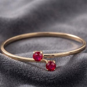 Puede incluir: Un brazalete de oro tipo bypass con dos rubíes rojos vibrantes de corte redondo, montados en engastes de cuatro garras en los extremos abiertos de la banda de oro pulido. El diseño presenta una estructura elegante con las gemas desplazadas.