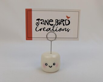 May include: A white, cylindrical marshmallow-shaped card holder with a smiling face, pink cheeks, and a thin silver metal wire loop on top. A rectangular white business card with black text reading Jane Bird Creations and a small orange bird icon.