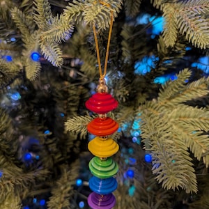 Könnte beinhalten: Ein vertikaler Hängeschmuck bestehend aus sechs gestapelten, scheibenförmigen Perlen in einer Regenbogenfarbfolge aus Rot, Orange, Gelb, Grün, Blau und Violett. Kleine goldfarbene Metallperlen trennen die Scheiben, befestigt an einer Goldschnur.