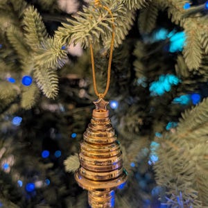 May include: A gold-colored, tiered, cone-shaped ornament with a metallic finish. The ornament features a small star at the top and is suspended by a thin gold cord. The structure consists of several stacked, circular, disc-like layers.
