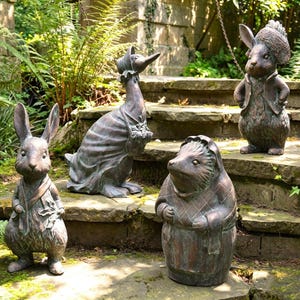 May include: Four bronze-toned garden statues featuring anthropomorphic animals: a rabbit wearing a scarf, a duck in a bonnet and shawl, a hedgehog in a quilted coat, and a rabbit wearing a jacket and hat, all standing on stone steps.