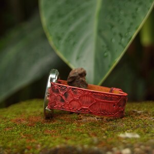 May include: A wristwatch featuring a vibrant red textured strap with a distinct scale-like pattern and visible stitching along the edges. The watch case is metallic silver and is attached to the strap, which displays a deep red hue with darker accents.