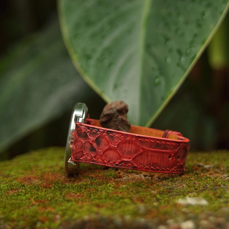 May include: A wristwatch featuring a vibrant red textured strap with a distinct scale-like pattern and visible stitching along the edges. The watch case is metallic silver and is attached to the strap, which displays a deep red hue with darker accents.