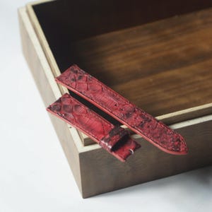 May include: A two-piece watch strap crafted from red leather with a textured, scale-like pattern. The strap features visible stitching along the edges, a tapered design, and a single fixed leather keeper loop on the shorter section of the band.